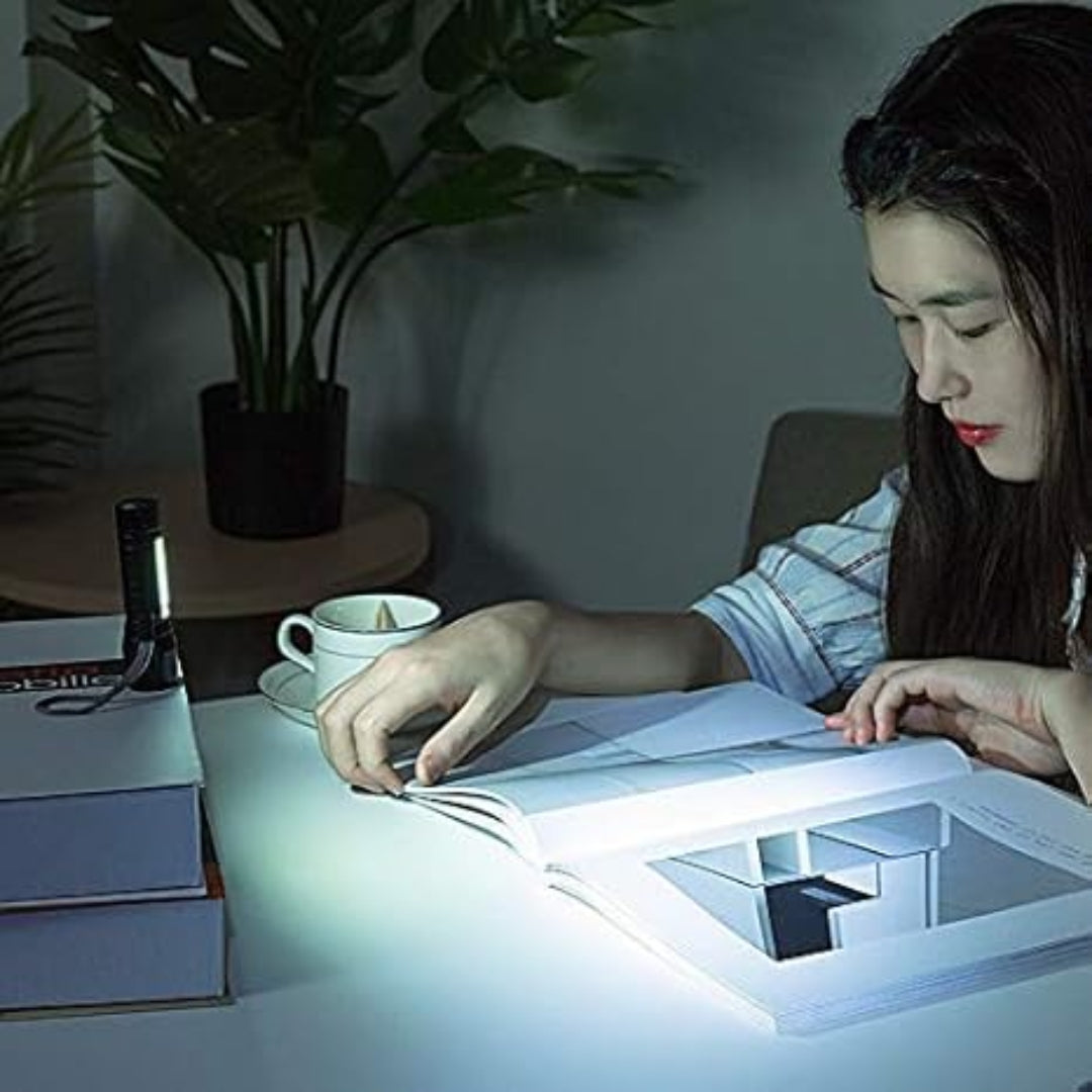 A woman reads a book at night illuminated by a compact flashlight placed on the desk beside her