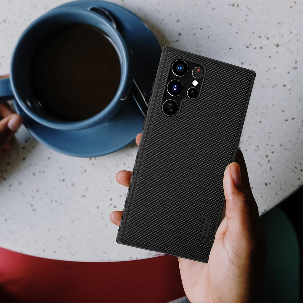 Hand holding a Samsung device in a matte black Nillkin mobile cover while having coffee, s22 ultra back cover reflecting a stylish and protective design.
